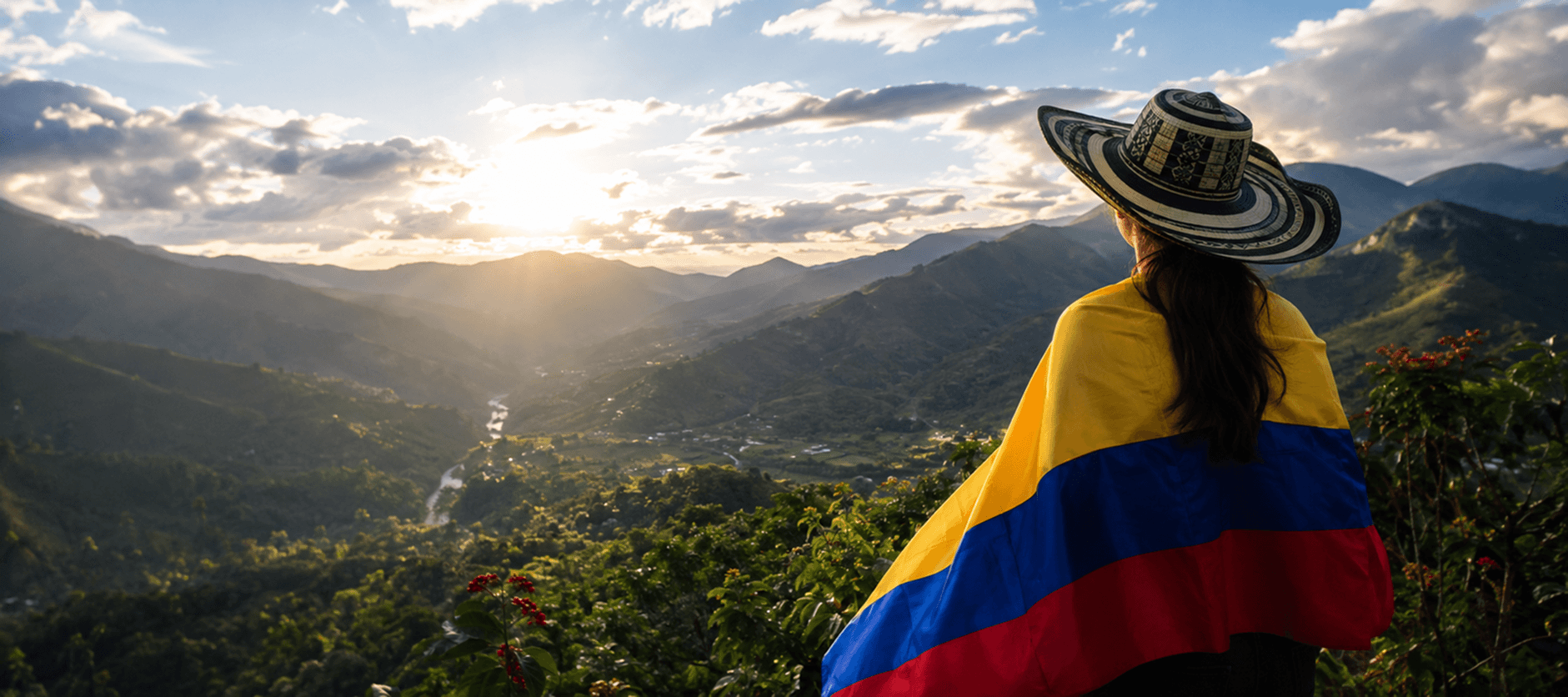 Una mujer con sombrero vueltiao, envuelta en la bandera de Colombia, mira el amanecer sobre un valle de montañas y cafetales.